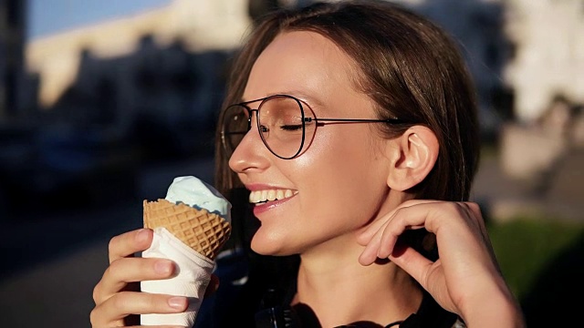 一个迷人的女孩在街上吃巨大的白色冰淇淋的侧面镜头。年轻女子戴着现代透明眼镜，微笑着，享受着她的时光