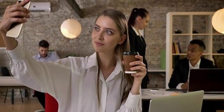 年轻迷人的女商人在办公室用智能手机端着咖啡自拍，她的同事们用科技、工作理念、商业理念、沟通理念进行网络交流