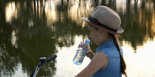 快乐的小女孩骑着自行车看着相机，微笑着在日落的河岸上。她戴着一顶帽子，站在以水为背景的地方，从一个清澈的瓶子里喝水。