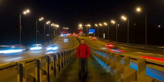 一个人站在公路上。随着时间的推移，高速公路的交通使汽车在夜间变得模糊不清。汽车在桥上行驶的晚上时间流逝。4 k UHD