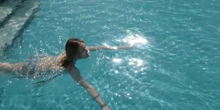 美丽的白人模特，年轻的女人是游泳在一个酒店的蓝色的水，在开放的天空下。放松、休假和自由的概念。慢动作