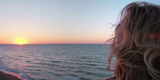 美丽的白种人卷发年轻女子日出或日落在海洋。风吹长了头发。她呼吸着海边的空气。自由的概念，生活的享受。慢动作