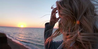 美丽的白种人卷发年轻女子日出或日落在海洋。风吹长了头发。她呼吸着海边的空气。自由的概念，生活的享受。缓慢的运动。近距离