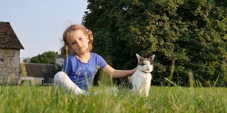 一个可爱的女孩和一只三色的猫坐在草地上。以一所老房子和几棵大树为背景
