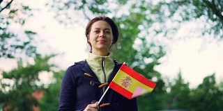慢动作肖像，漂亮的年轻女子西班牙球迷挥舞西班牙官方旗帜，看着镜头与美好的微笑。背景是绿树和天空。