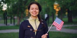 慢动作肖像的骄傲的美国公民微笑妇女挥舞美国国旗，看着相机和微笑。公园里的绿树、草坪、小路都看得见。