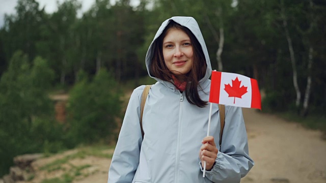 慢动作肖像的女旅行者漂亮的女孩拿着加拿大国旗，微笑着看着相机与美丽的自然景观在背景。