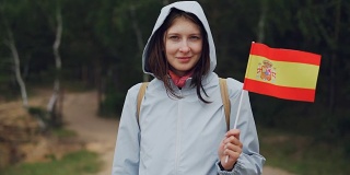 慢动作肖像的快乐世界旅行者漂亮的女孩在帽外套挥舞西班牙国旗和看着镜头微笑。国家和旅游概念。