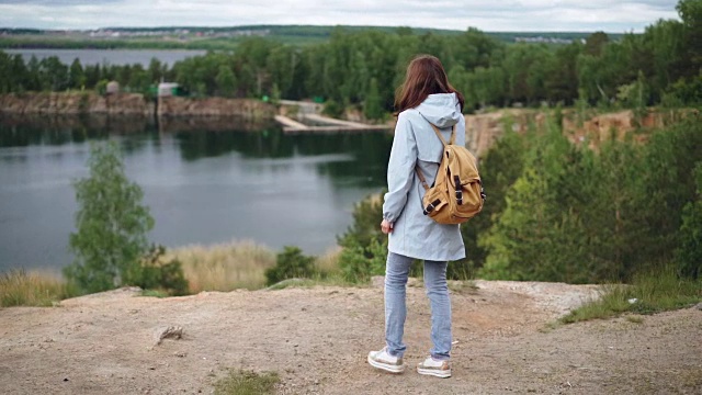 一名年轻的红发女子走到悬崖边，观看令人兴奋的湖泊、树林和建筑物的慢动作肖像。美丽的自然和旅游理念。