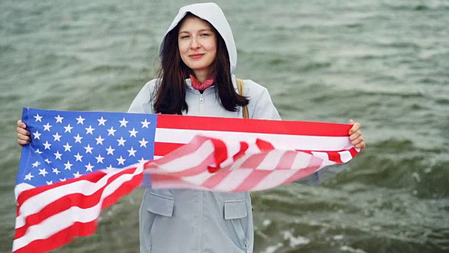 慢动作肖像快乐的美国女孩举着美国国旗和移动它站在海岸边与波浪可见。国籍和国家概念。