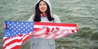 慢动作肖像快乐的美国女孩举着美国国旗和移动它站在海岸边与波浪可见。国籍和国家概念。