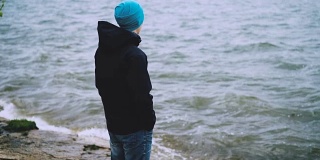 慢动作肖像体贴的家伙在帽子，夹克和牛仔裤站在海边，看着海浪享受自然。健康的生活方式和旅游理念。