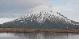 游客漫步在新西兰北岛的塔拉纳基火山，与山和登山者在湖中的倒影在一起。新西兰被