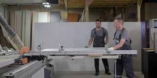 家具制造车间，两名工人正在把刨花板放到切割机上