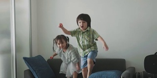 泰国可爱的小女孩和蹒跚学步的男孩在客厅的沙发上跳舞和玩耍-库存视频