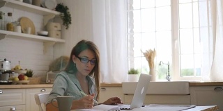 年轻的自由职业女性，戴着眼镜在笔记本电脑上工作，在明亮的厨房里做笔记
