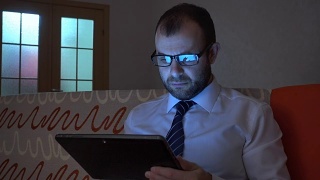 一个穿着白衬衫，系着蓝领带的严肃男人晚上在家用平板电脑工作。商人在平板电脑上工作，晚上坐在沙发上。视频素材模板下载