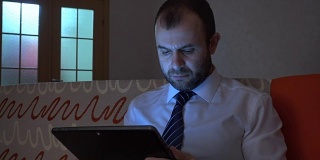 一个穿着白衬衫，系着蓝领带的严肃男人晚上在家用平板电脑工作。商人在平板电脑上工作，晚上坐在沙发上。