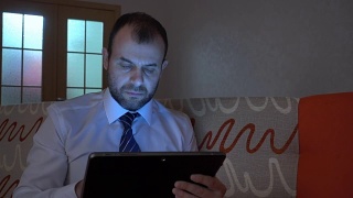 一个穿着白衬衫，系着蓝领带的严肃男人晚上在家用平板电脑工作。商人在平板电脑上工作，晚上坐在沙发上。视频素材模板下载