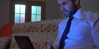穿着白衬衫，系着蓝领带的严肃男士晚上在家用笔记本电脑工作。在笔记本上工作，晚上坐在沙发上。