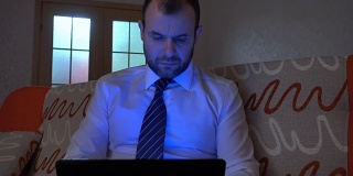穿着白衬衫，系着蓝领带的严肃男士晚上在家用笔记本电脑工作。在笔记本上工作，晚上坐在沙发上。
