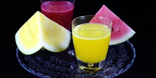 金字塔状的红黄西瓜和几杯鲜榨的西瓜汁，在黑色背景下对着时钟旋转近距离。