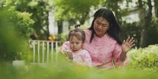一位泰国家长教她的女儿唱歌和跳舞，同时使用智能手机在花园里放松