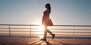 穿着复古短裙的年轻女子踏在海边的木堤岸上。女孩在日出或日落的背景下行走。美丽的女性腿的场景。慢动作