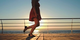穿着复古短裙的年轻女子在海边的木制堤岸上奔跑。女孩在日出或日落的背景下行走。美丽的女性腿的场景。慢动作