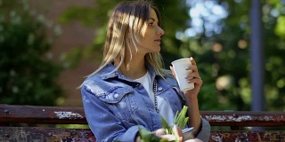 美丽的金发美女穿着牛仔夹克放松在公园长椅上喝咖啡