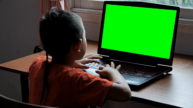 亚洲男孩7岁用笔记本电脑在家里搜索互联网信息的绿色屏幕。日常生活中的技术概念。