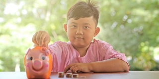 亚洲男孩带着快乐的微笑把硬币放入储蓄罐，慢镜头。散景背景。存钱，为将来攒钱。