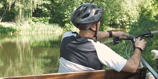 一个人骑着自行车，在公园里夏天休息。他坐在池塘边的长椅上。