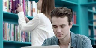 男生在大学图书馆专注地用笔记本电脑做作业，女生在后台浏览书架上的书