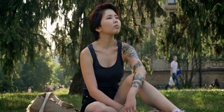 时髦的年轻亚洲女性潮人与纹身坐在草地上附近的大学，深思