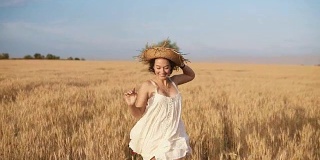 穿着白衣服的漂亮年轻女子在麦田里奔跑。微笑着，快乐的女性跑着，拿着她的草帽在她的头上。前视图