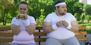 肥胖的年轻夫妇吃汉堡，沉迷于垃圾食品，缺乏意志力