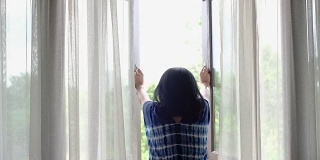 亚洲妇女在早晨打开窗帘和窗户看外面的阳台度假。暑假假期