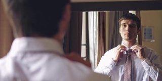 一名穿着白衬衫的英俊男子站在镜子前系着领带。将目光从人转向镜子。3840 x2160