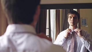 一名穿着白衬衫的英俊男子站在镜子前系着领带。将目光从人转向镜子。3840 x2160视频素材模板下载