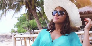 快乐美丽的女人戴着帽子和太阳镜度假，坐在海滩咖啡馆与风景的大海和棕榈树喝茶，吃早餐。缓慢的运动。3840 x2160