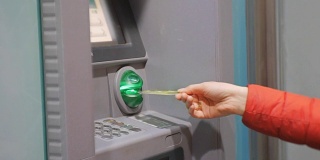 使用自动提款机的女人。身着红色泡泡夹克的女性手将信用卡插入ATM机并在晚上输入密码。