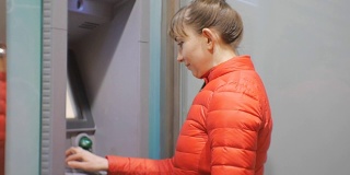 使用自动提款机的女人。一位穿着红色泡泡夹克的年轻美女将信用卡插入ATM机并输入密码。