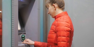 使用自动提款机的女人。穿着红色泡泡夹克的年轻美女正在将信用卡插入自动取款机。女人微笑着输入密码。