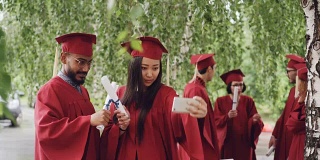 同学们微笑着摆着毕业证书自拍，女孩拿着智能手机，人们穿着长袍和帽子。教育与现代生活理念。