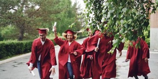 慢镜头中，快乐的毕业生们穿着红袍和红帽，挥舞着毕业证书，微笑着在校园里奔跑。美丽的树木和灌木可见，下雨了。