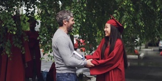 一位慈爱的父亲在毕业典礼上祝贺他的女儿，人们在户外拥抱和欢笑，背景中可以看到其他毕业生。