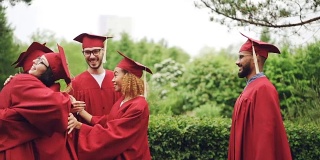 慢镜头中，欢乐的女生和男生们戴着学士帽和学士服，拥抱握手，在户外享受毕业的日子。
