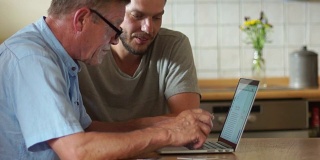 一个成熟的男人和他已经成年的儿子在电脑旁边。帮助退休人员，网上购物，父亲节